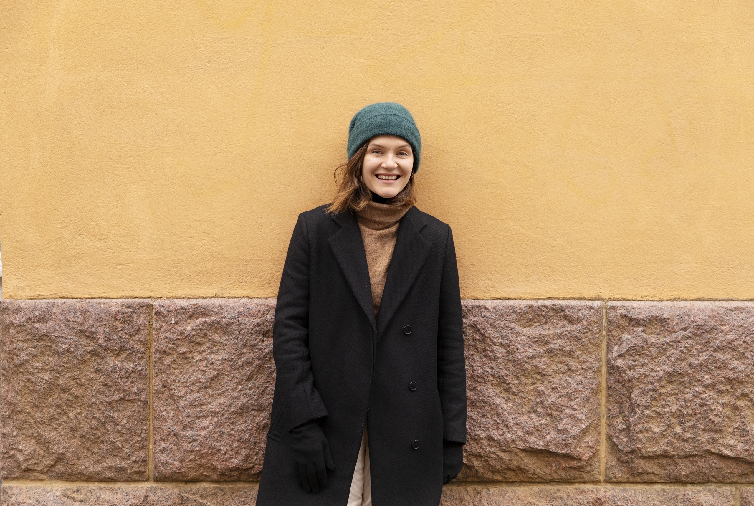 Kuvassa Ronja Salmi nojaa oranssiin seinään ja katsoo kameraan hymyillen. Hänellä on vihreä pipo ja musta takki.