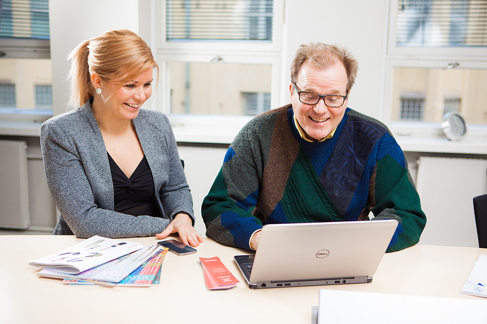 Colleagues Juha Kallanranta and Meri Saarlinna are smiling and sitting by the table with one laptop and some broschures