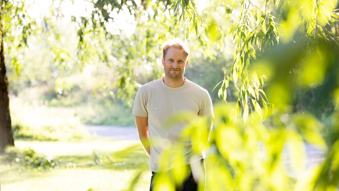 Kuvittaja Antti Kalevi on kuvan keskellä, seisoo, hymyilee ja katsoo kameraan. Hän on ulkona, lehtipuiden ympäröimänä ja aurinko paistaa.