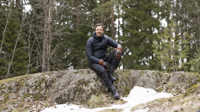 Peltola on metsän keskellä, istuu kalliolla, katsoo kameraan ja hymyilee.