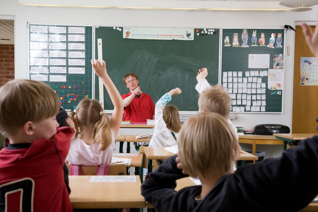 Luokkahuone alakoulussa. Lapset viittaavat ja opettaja osoittaa sormella.