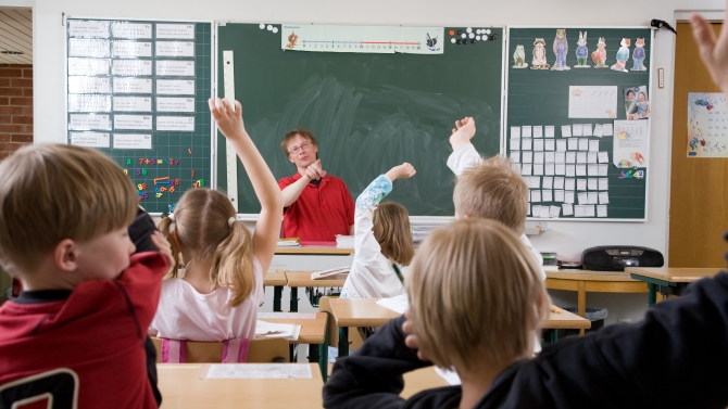 Luokkahuone alakoulussa. Lapset viittaavat ja opettaja osoittaa sormella.